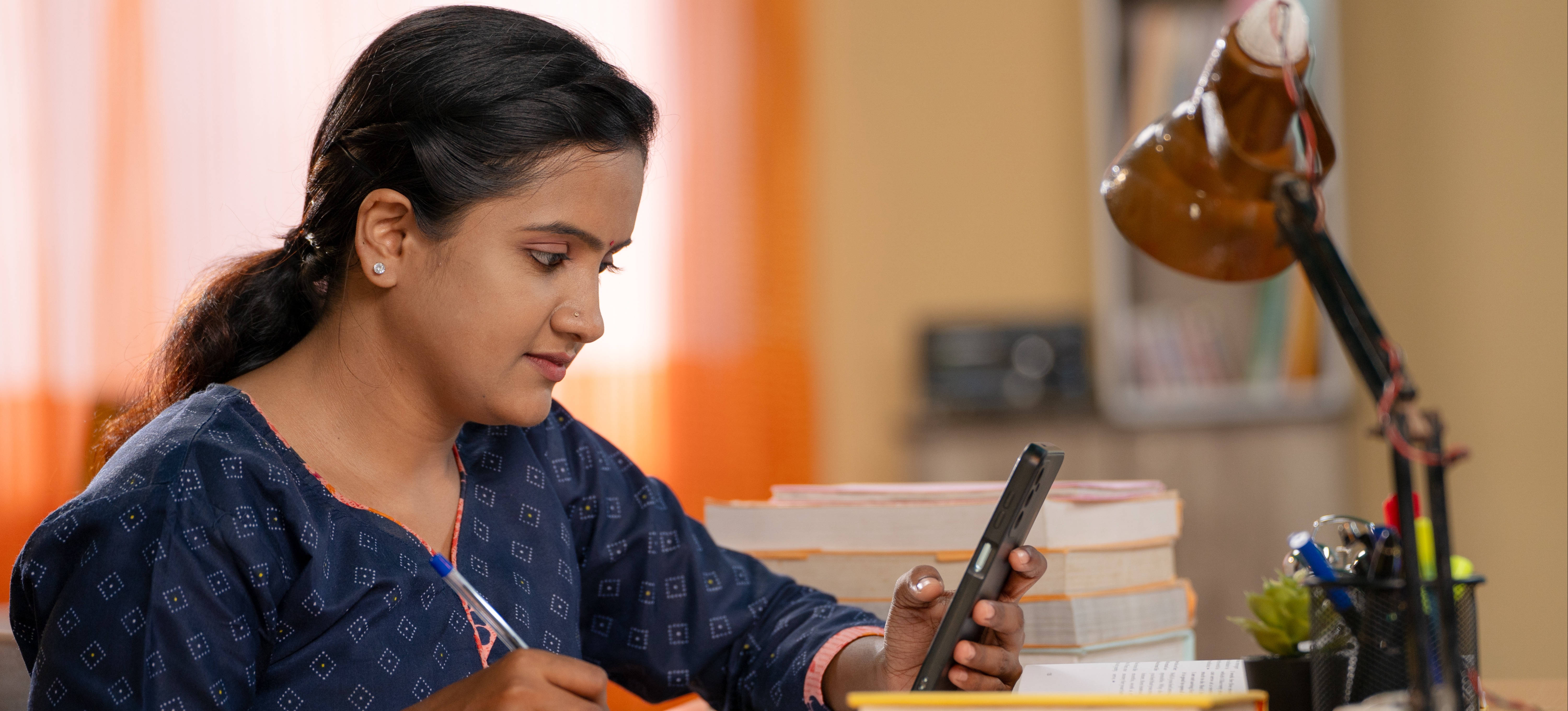 [Featured Image] An aspiring young doctor uses her smartphone and books to study for the NEET exam.  