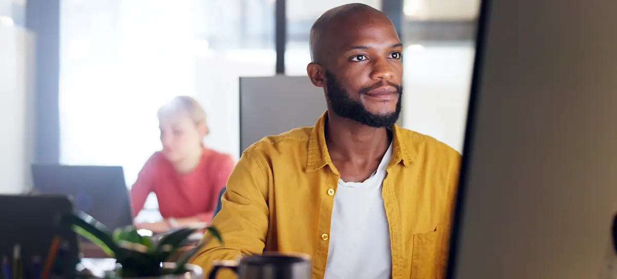 [Featured Image] A service desk analyst sits at a desk looking at their computer while a colleague works at their computer in the background.