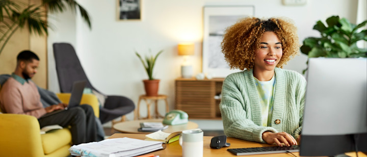 [Featured Image] Two people work on computers, one on a laptop sitting on a couch in the background and the other on a desktop in the foreground, transmitting data online thanks in part to the transport layer.
