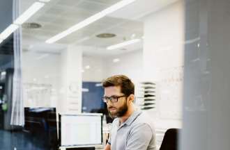 [Featured Image]: A male, site reliability engineer, wearing a gray shirt, and glasses, sitting in front of his desktop, in his office. 