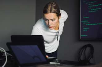 [Featured image] A cybersecurity analyst in a white shirt leans over their desk to view their laptop screen. They are in a dark office and a large monitor with green code is on the wall behind them.