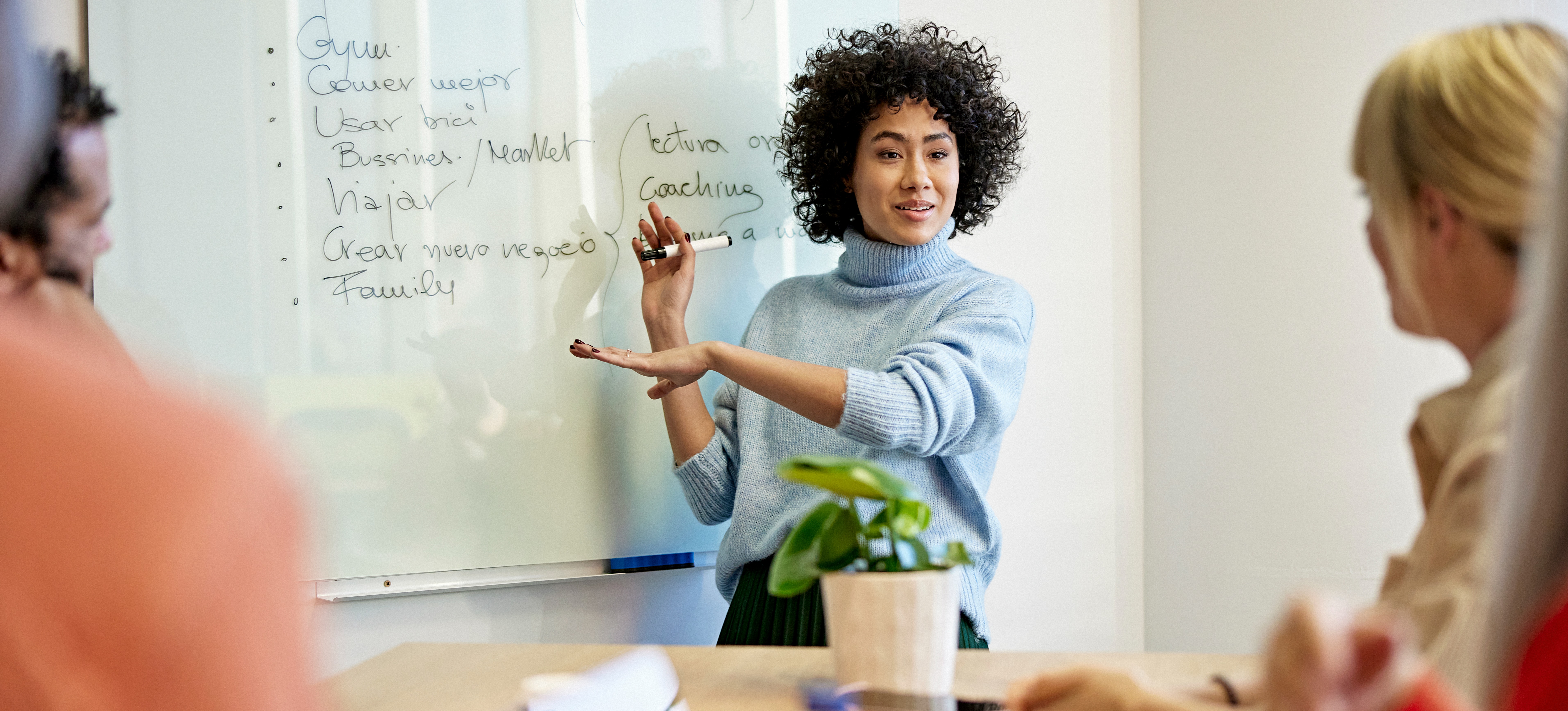 [Featured Image] Woman in her late 20s standing in front of a whiteboard using her coaching skills to address her colleagues in a meeting room.