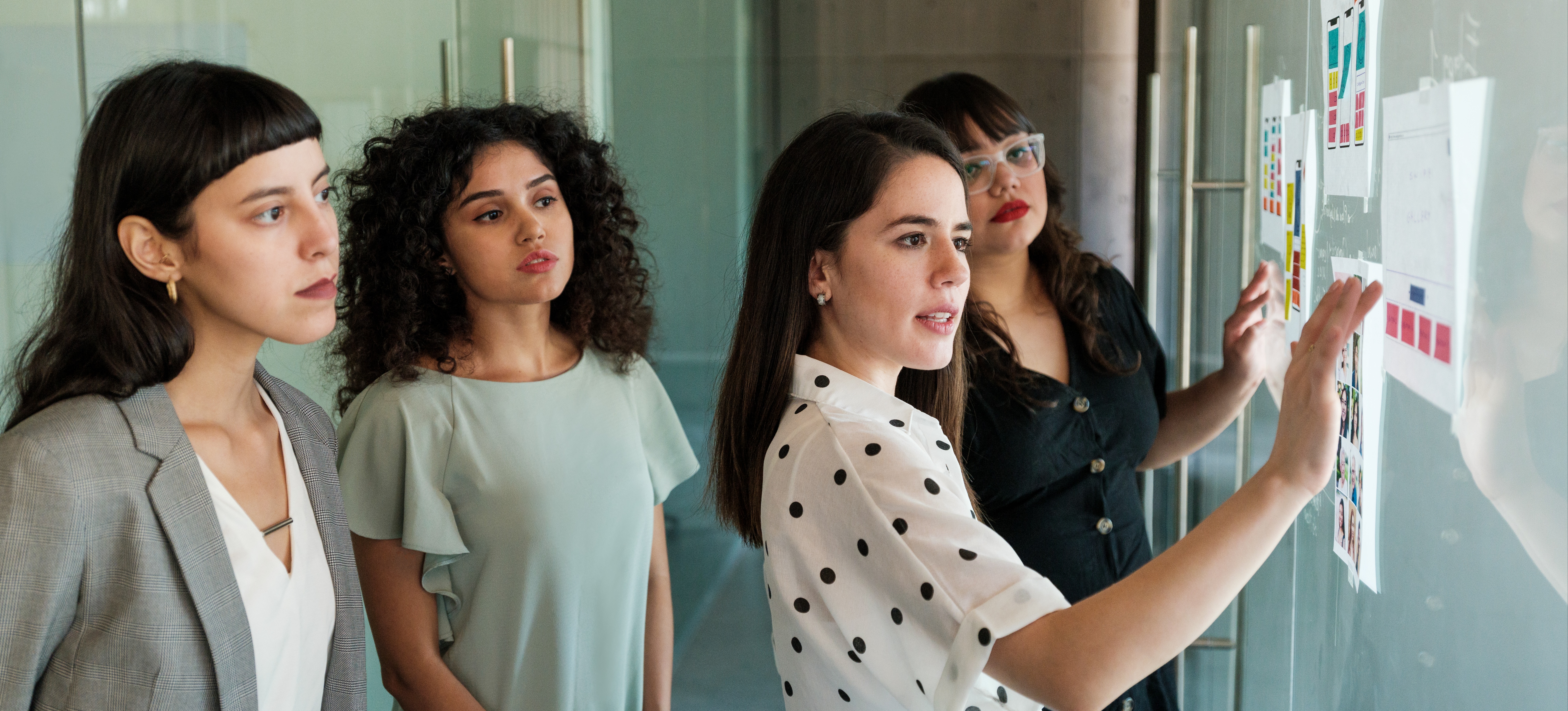 [Hauptbild] Vier Geschäftsfrauen stehen um ein Whiteboard herum, betrachten die angehängten Zettel und diskutieren den UX-Designprozess.