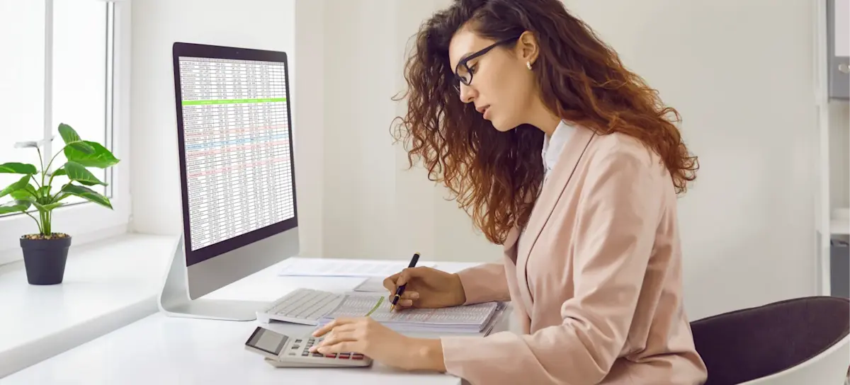 [Featured Image] An accountant who has earned an online accounting degree sits at a computer in a sunny office, viewing a spreadsheet and working with a calculator.
