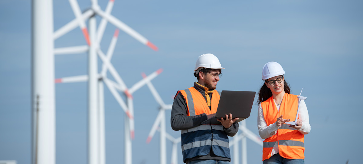 [Featured Image] Two wind turbine technicians, one of the highest paying jobs in energy, walk through a wind farm, with the wind turbines behind them, while wearing protective gear.
