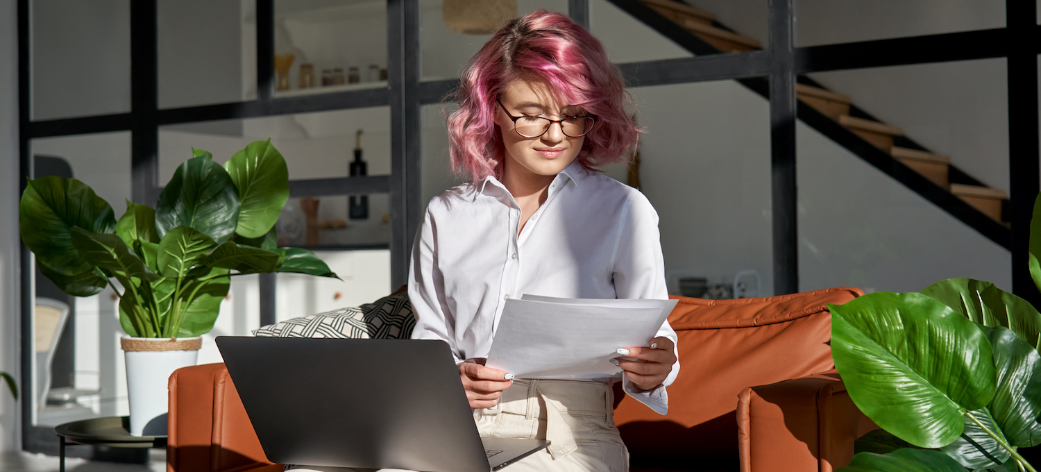 [Featured image] A person with pink hair and glasses sits on an orange sofa. They glance at two pieces of paper in their hand. 