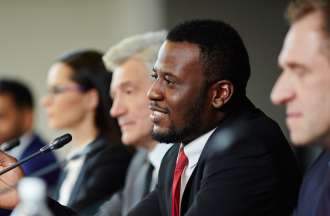 [Featured Image]: Man wearing a dark suit, red tie, and white shirt leading a panel. The panel includes three men and one woman.