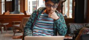 Featured image: A man wearing a blue striped shirt and glasses sits in a cafe and reads a job positing on his computer. 