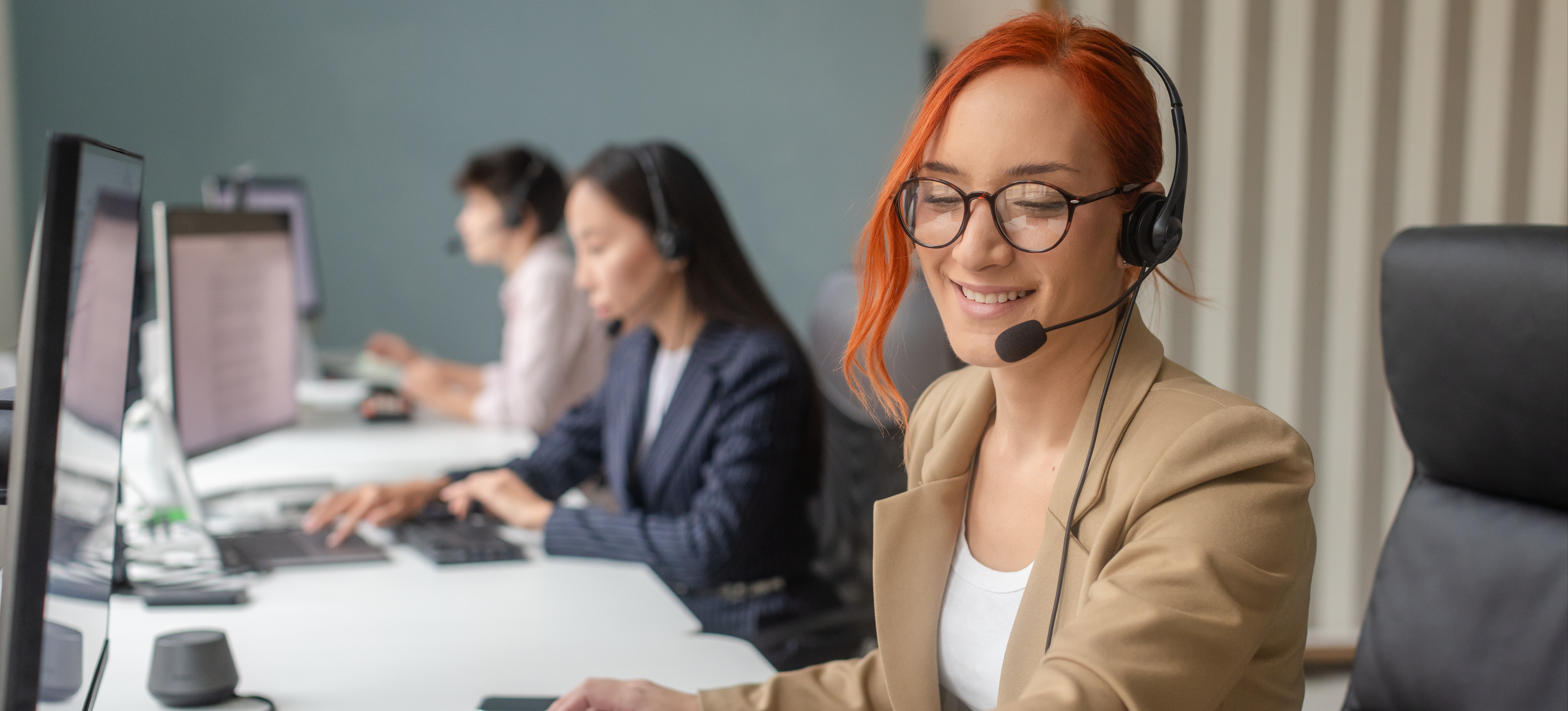 [Featured Image] A team of three professionals is sitting at a long desk in front of their computers, working efficiently. The person in the foreground is smiling and interacting with a client on a tablet to the left of their computer. 
