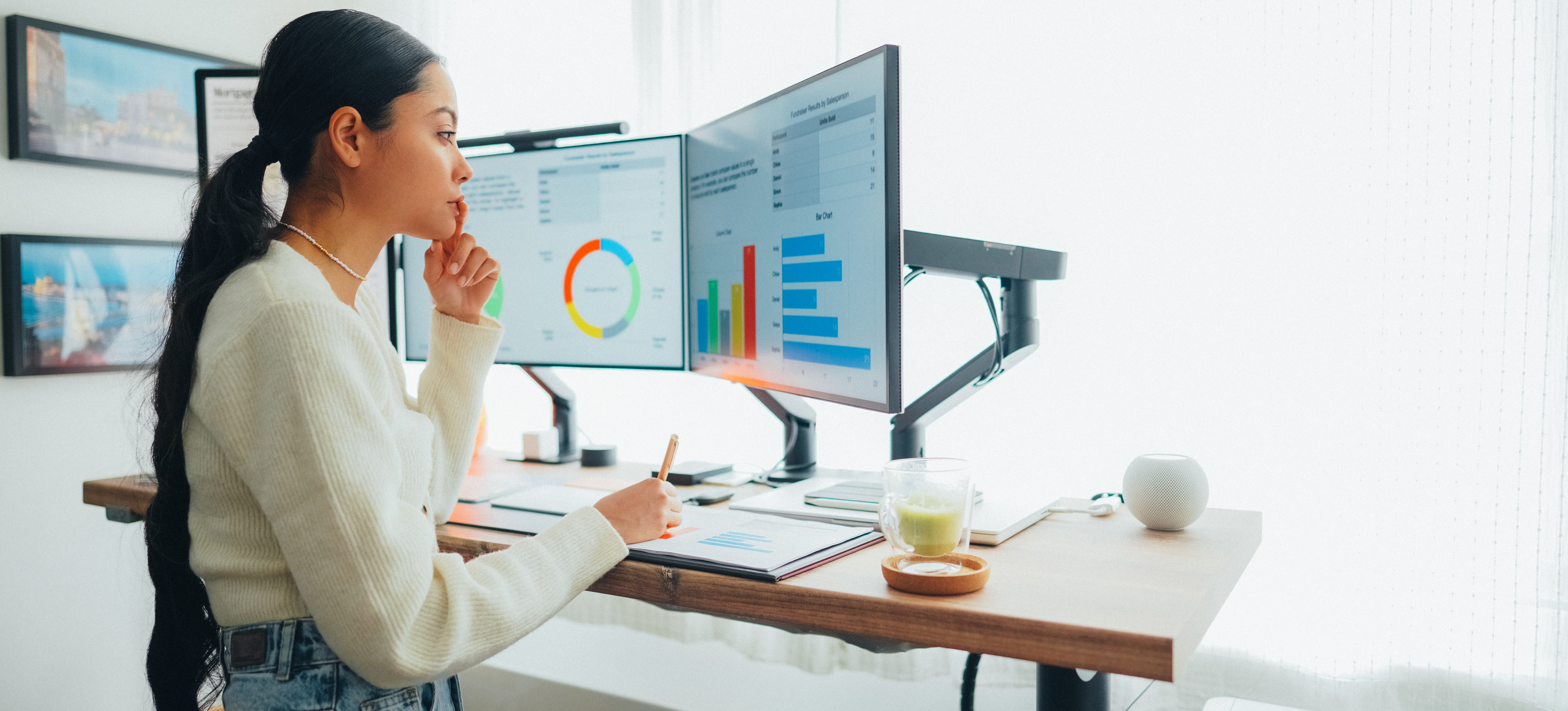 [Featured Image] An IT business analyst stands at her office desk looking at dual monitors that display data and graphs
