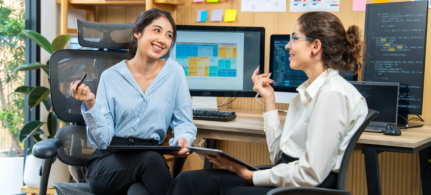 [Featured Image] Two rofessionals discuss Perplexity AI and if it’s the right choice for their project while holding tablets, with computers displaying AI platforms in the background.
