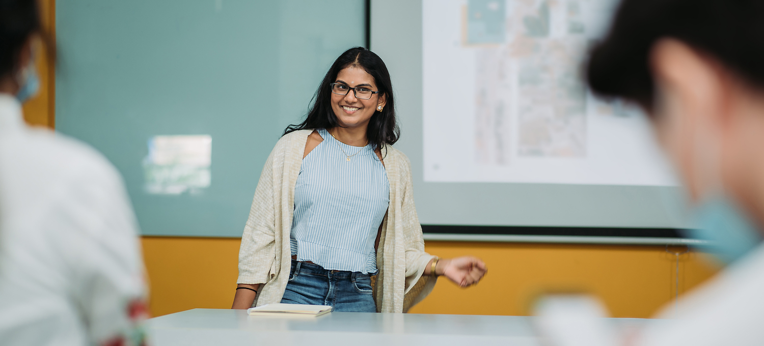 [Featured image] A young person with dark hair and glasses stands giving a presentation.