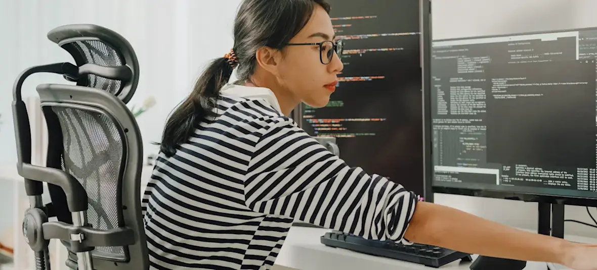 [Featured Image] A data scientist wearing glasses and a striped shirt working in front of several computer monitors. 
