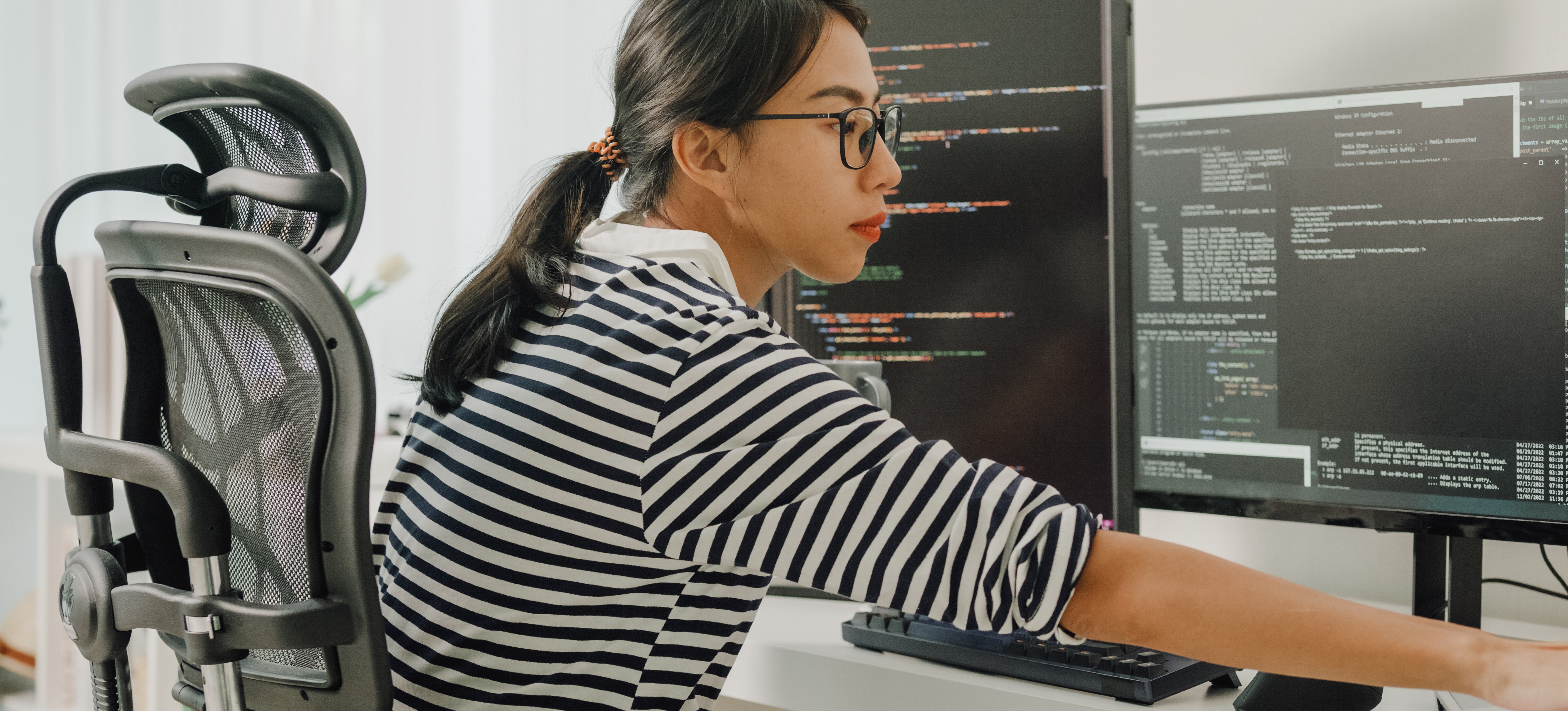 [Featured Image] A data scientist wearing glasses and a striped shirt working in front of several computer monitors. 
