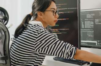 [Featured Image] A data scientist wearing glasses and a striped shirt working in front of several computer monitors. 
