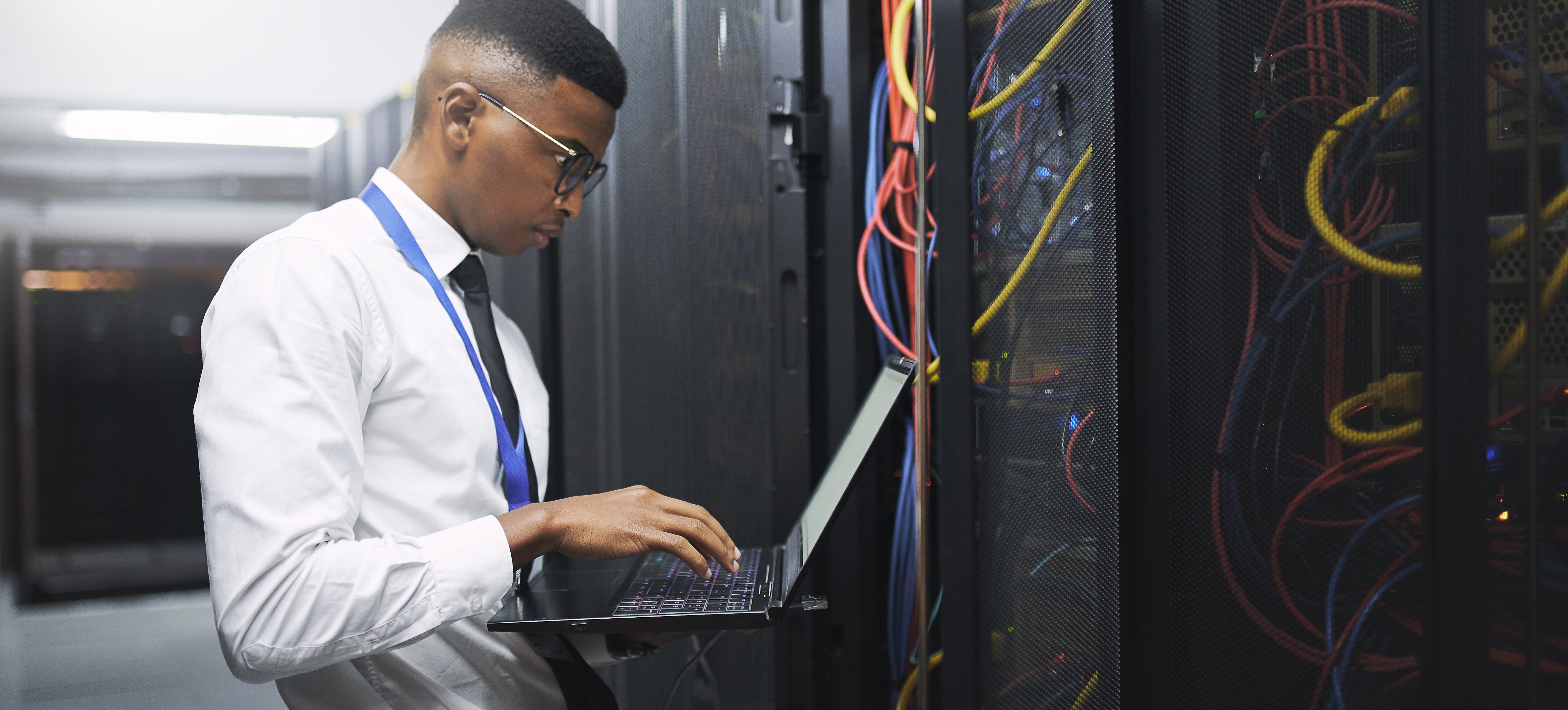 [Featured Image] A system administrator degree-holder stands in a server room holding a laptop and assessing the server equipment.
