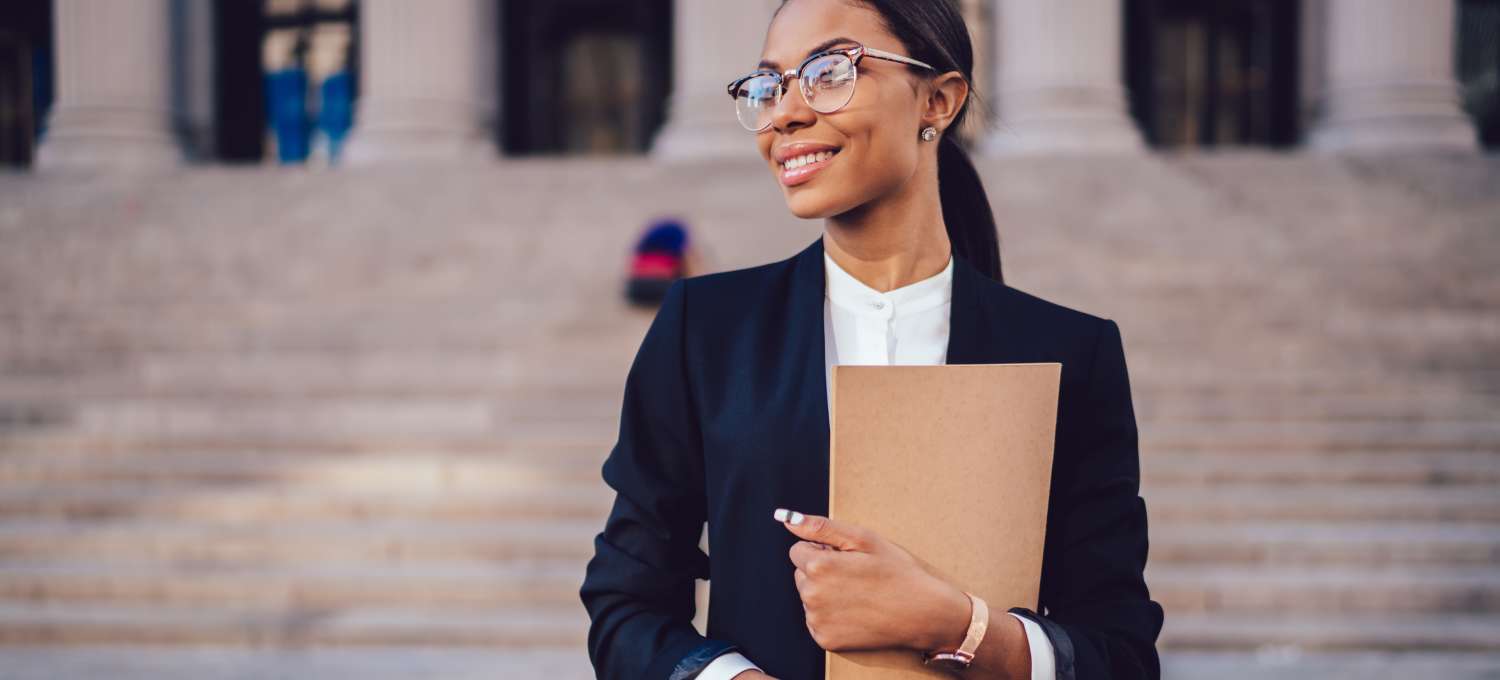 [Image] A woman wearing glasses and a black suit holds a tan folder.