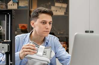 [Featured image] A computer programmer is working on a laptop and holding a VR set. 