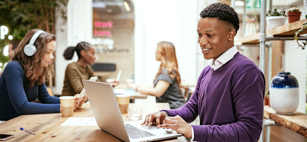 [Featured Image] A person with a marketing degree wearing a purple sweater works on their laptop at a shared working station. Three people sit in the background. 