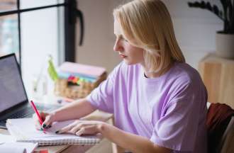 [Featured image] A blonde woman in a pink t-shirt sits at a desk writing in a notebook.