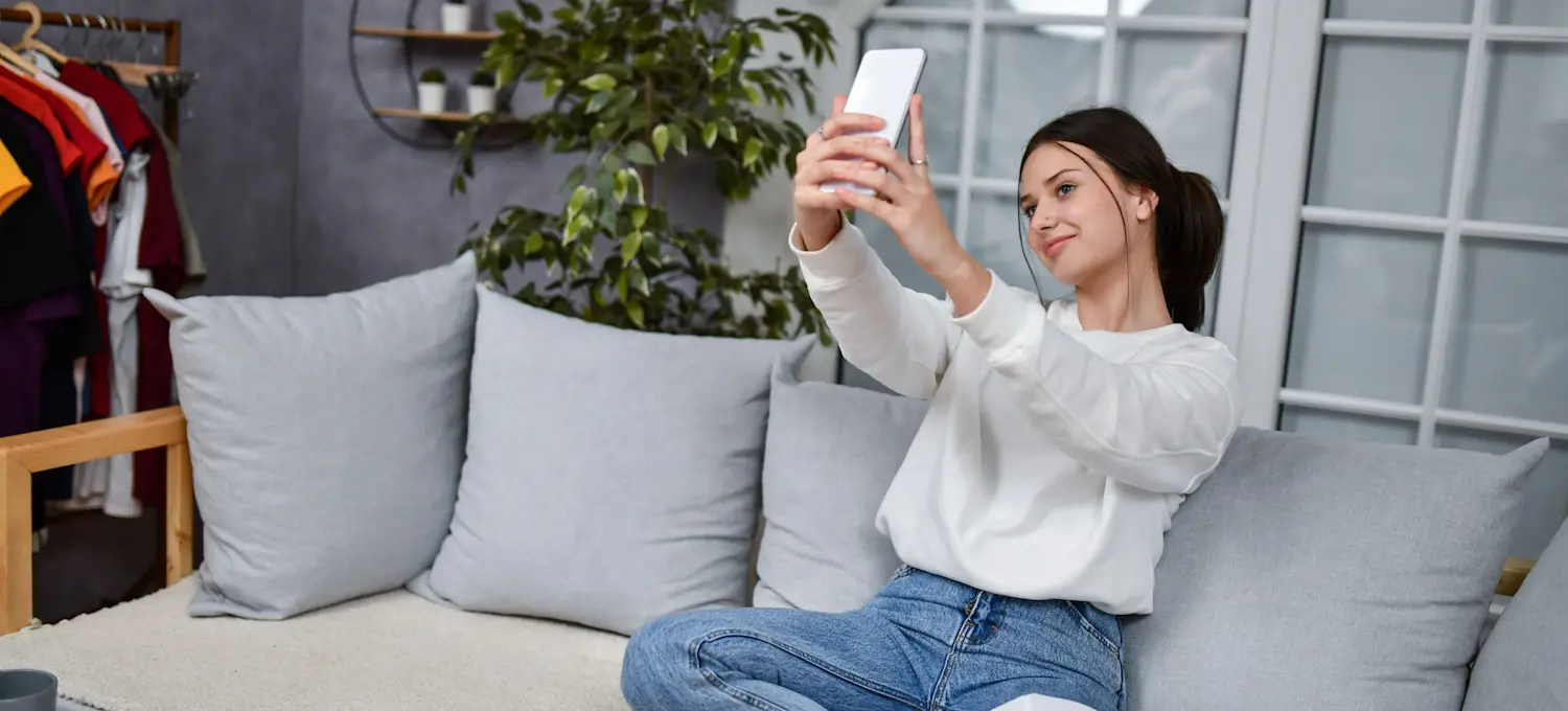 [Featured Image] A person sitting on a couch in their living room holds up their smartphone at arm's length to use an AR filter.
