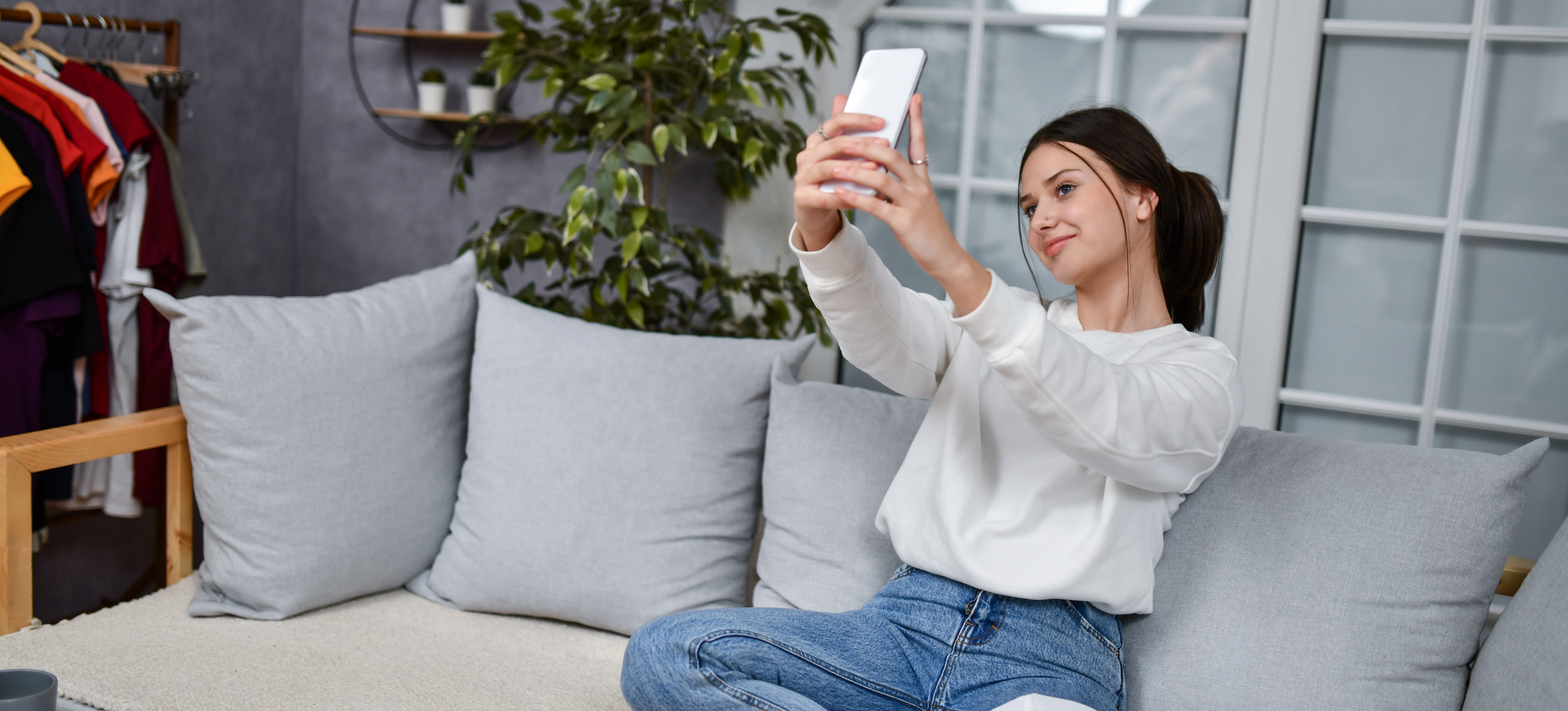 [Featured Image] A woman sitting on a couch in her living room holds up her smartphone at arm's length to use an AR filter.
