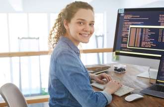 [Featured image] A young person with long curly hair in a ponytail sits in front of a desktop and laptop. They're turned, smiling at the camera. 