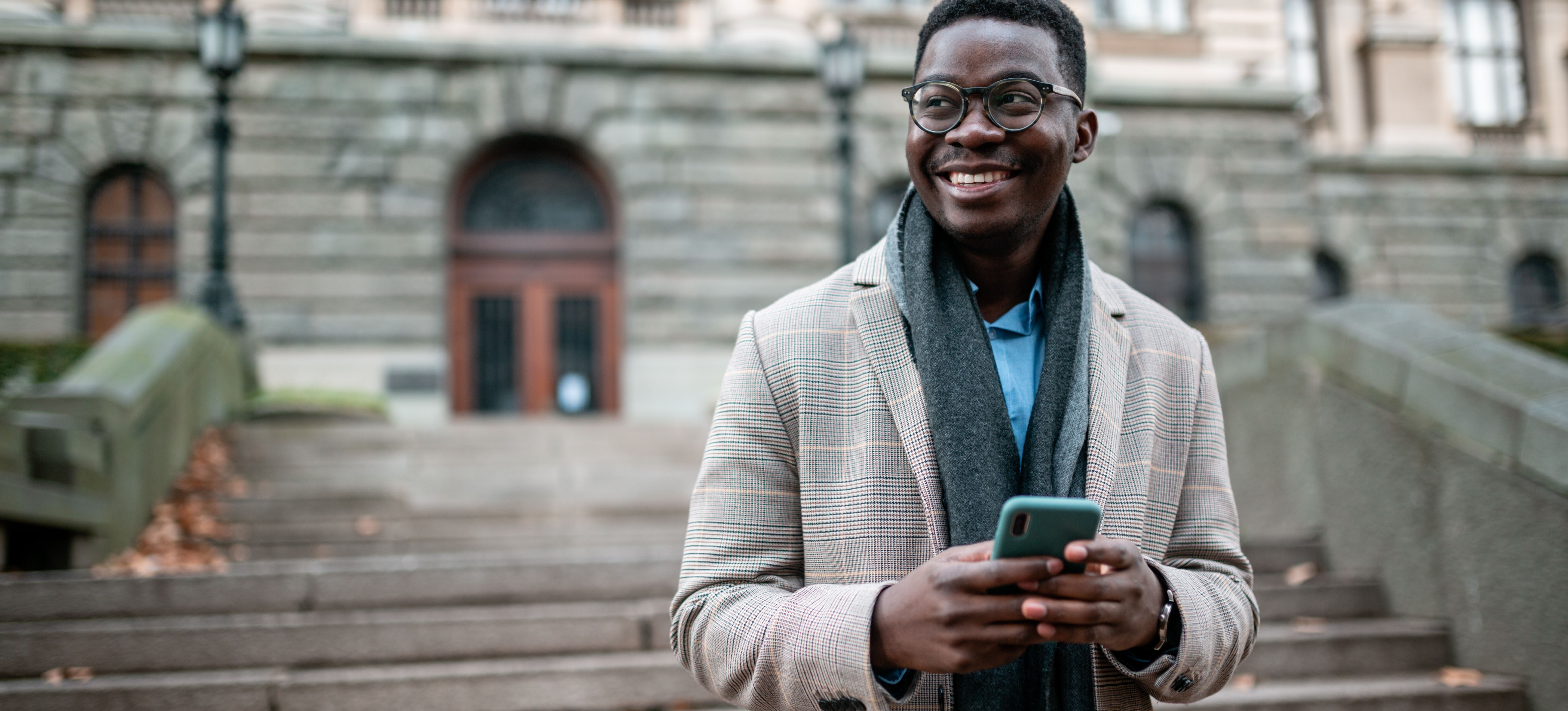 [Featured image] A Doctorate of Business Administration student stands on the steps of a university building while using their cell phone. They're wearing a jacket and scarf.