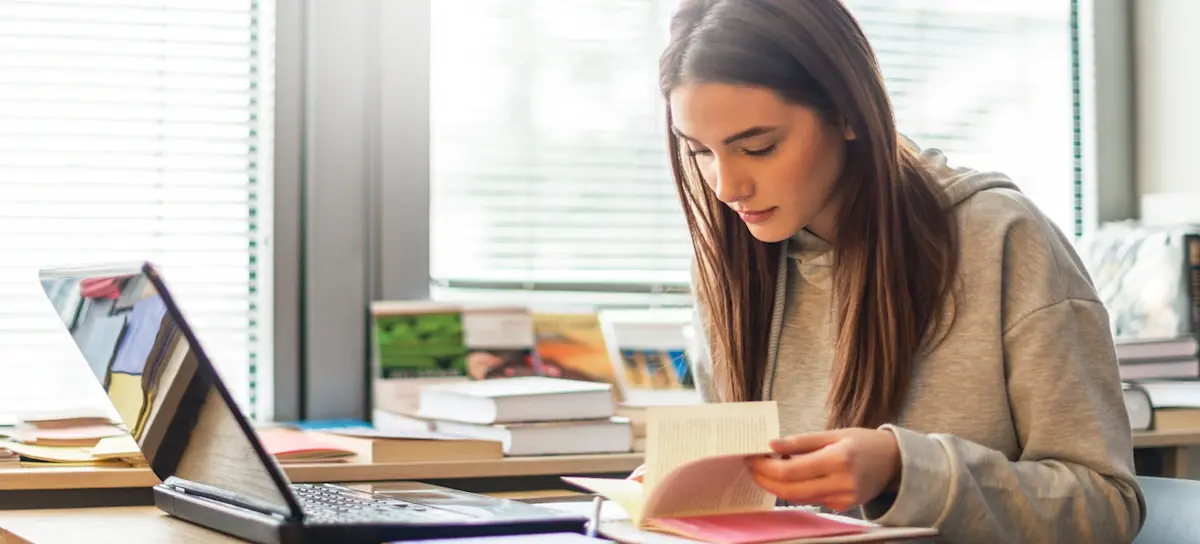 [Featured Image] A person sits with an open book in front of their laptop while studying for a bachelor's degree. 