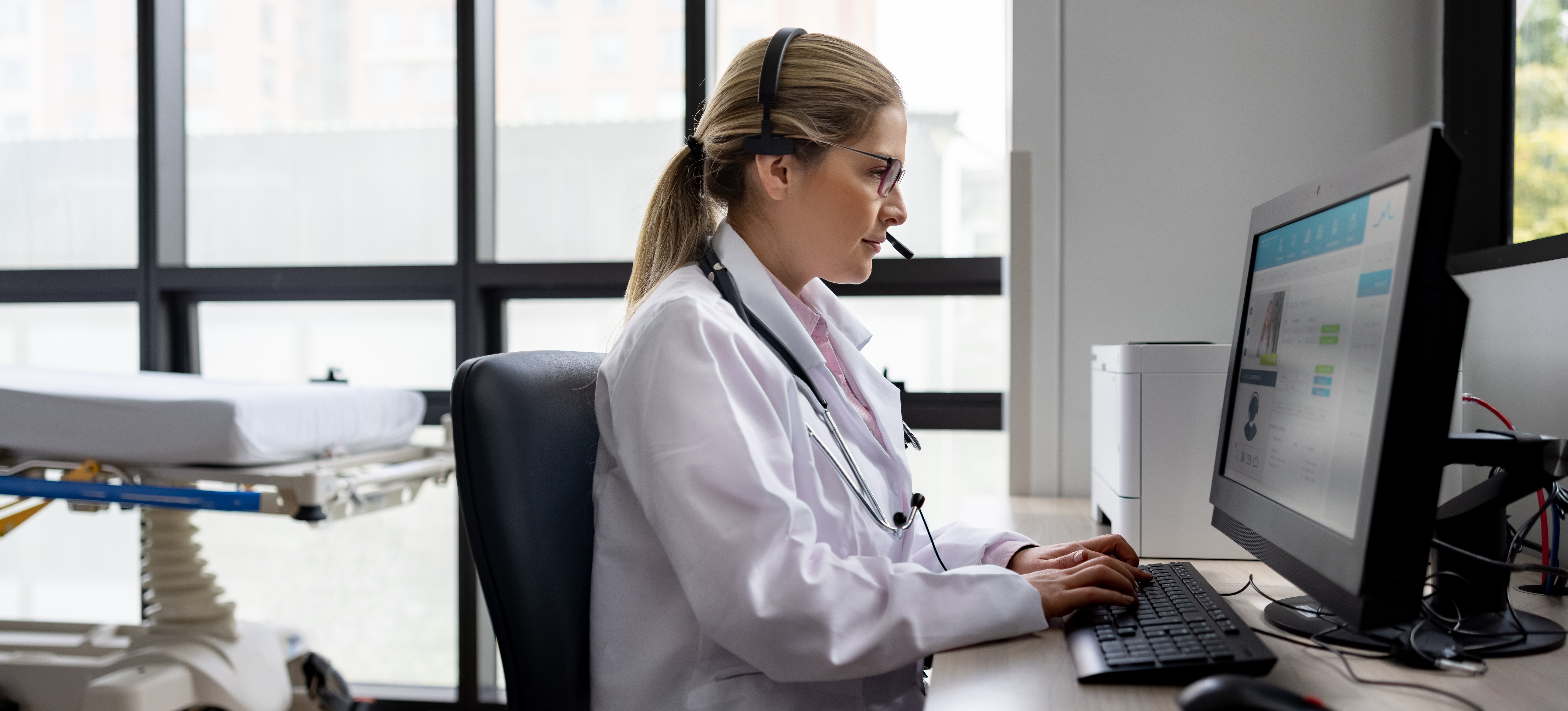 [Featured Image] A Medical Transcriptionist in a medical office sits at a desktop computer and types.