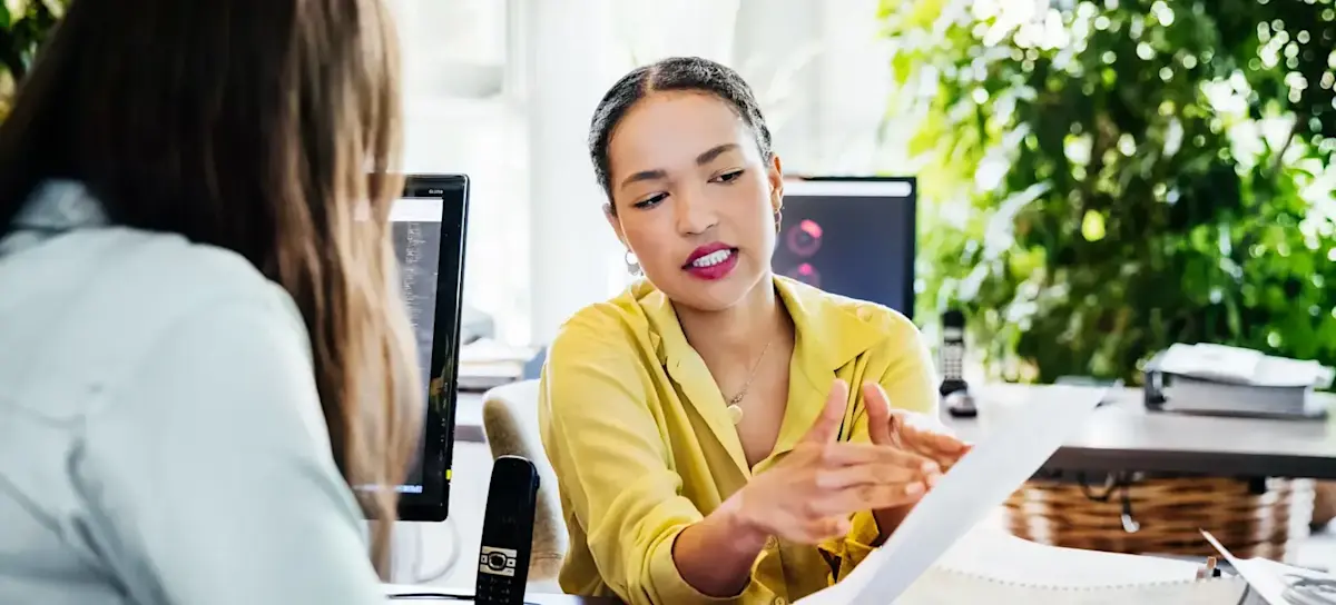 [Featured Image]: A business analyst reviews data with a client while sitting at a desk.
