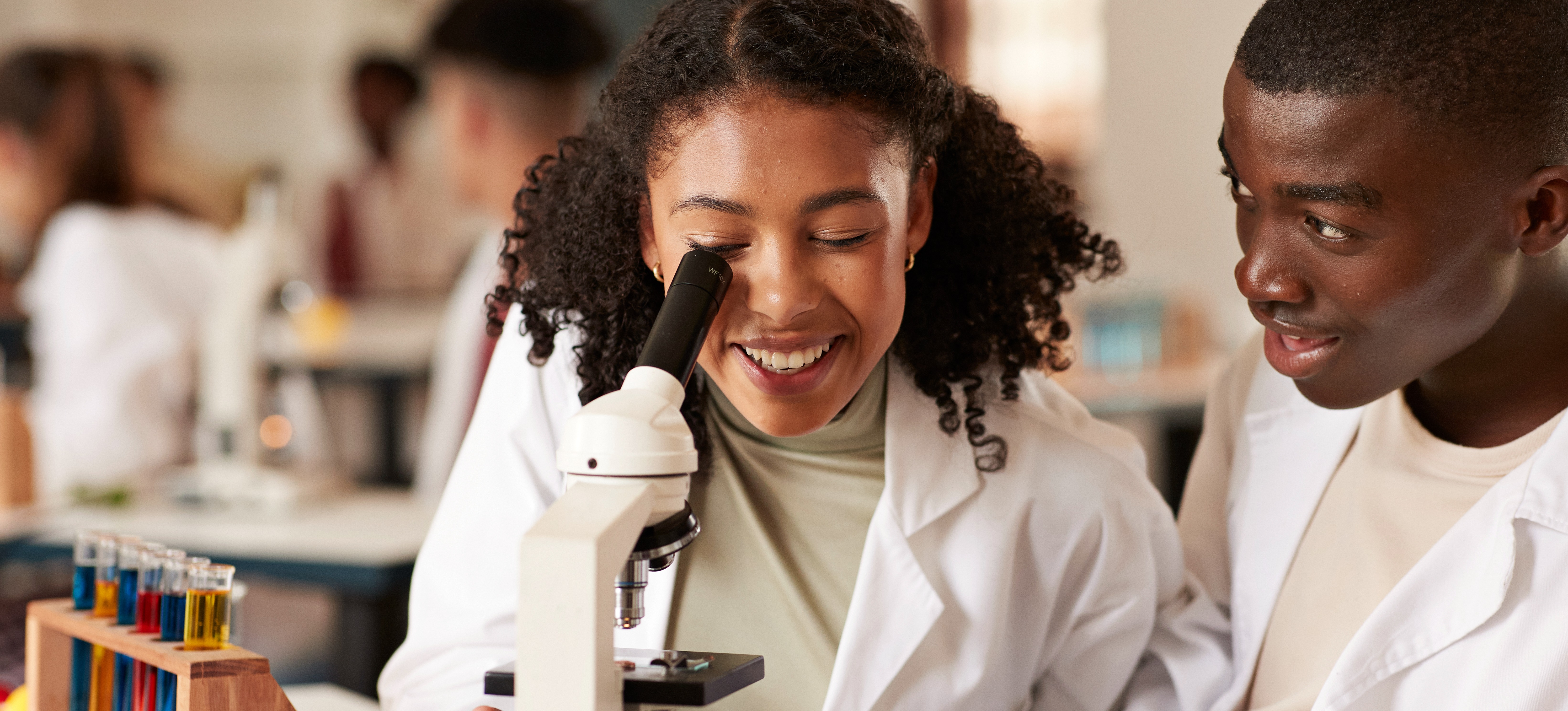 [Featured Image] Two students look at a microscope in a classroom while earning an undergrad degree for med school.   