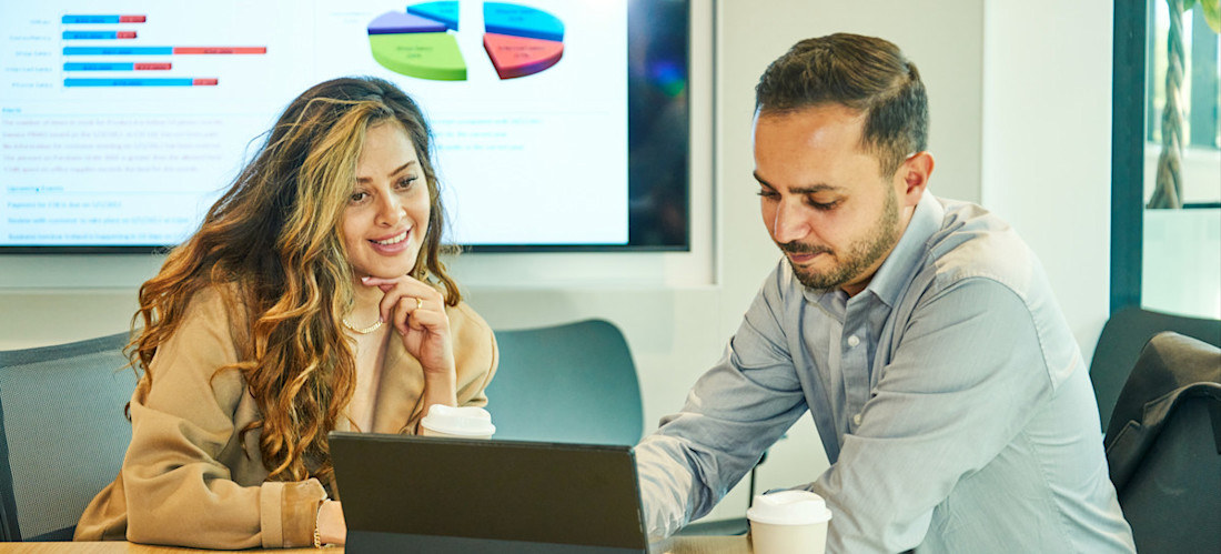 [Featured Image] A business intelligence developer sits in a conference room with a laptop and discusses data with a colleague.