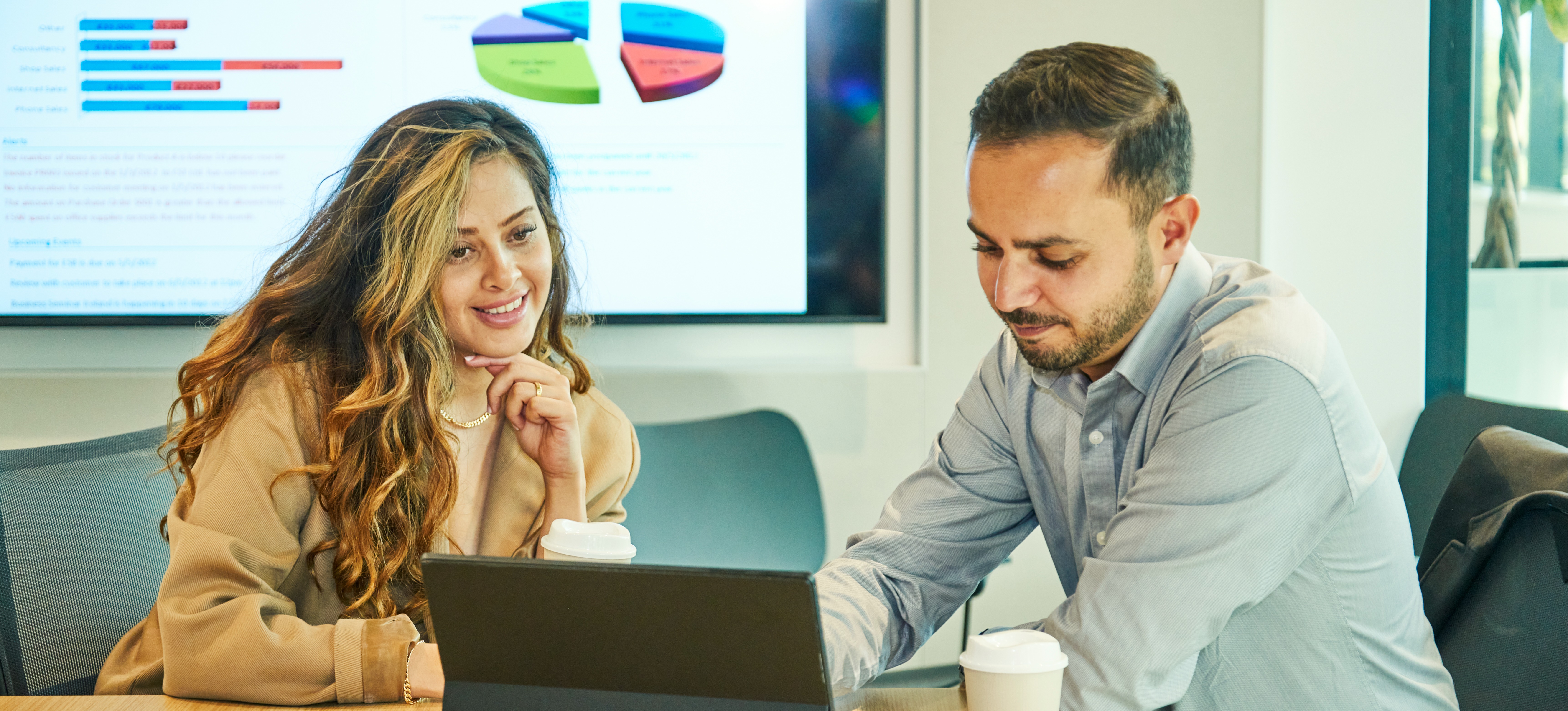 [Featured Image] A business intelligence developer sits in a conference room with a laptop and discusses data with a colleague.