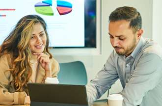 [Featured Image] A business intelligence developer sits in a conference room with a laptop and discusses data with a colleague.