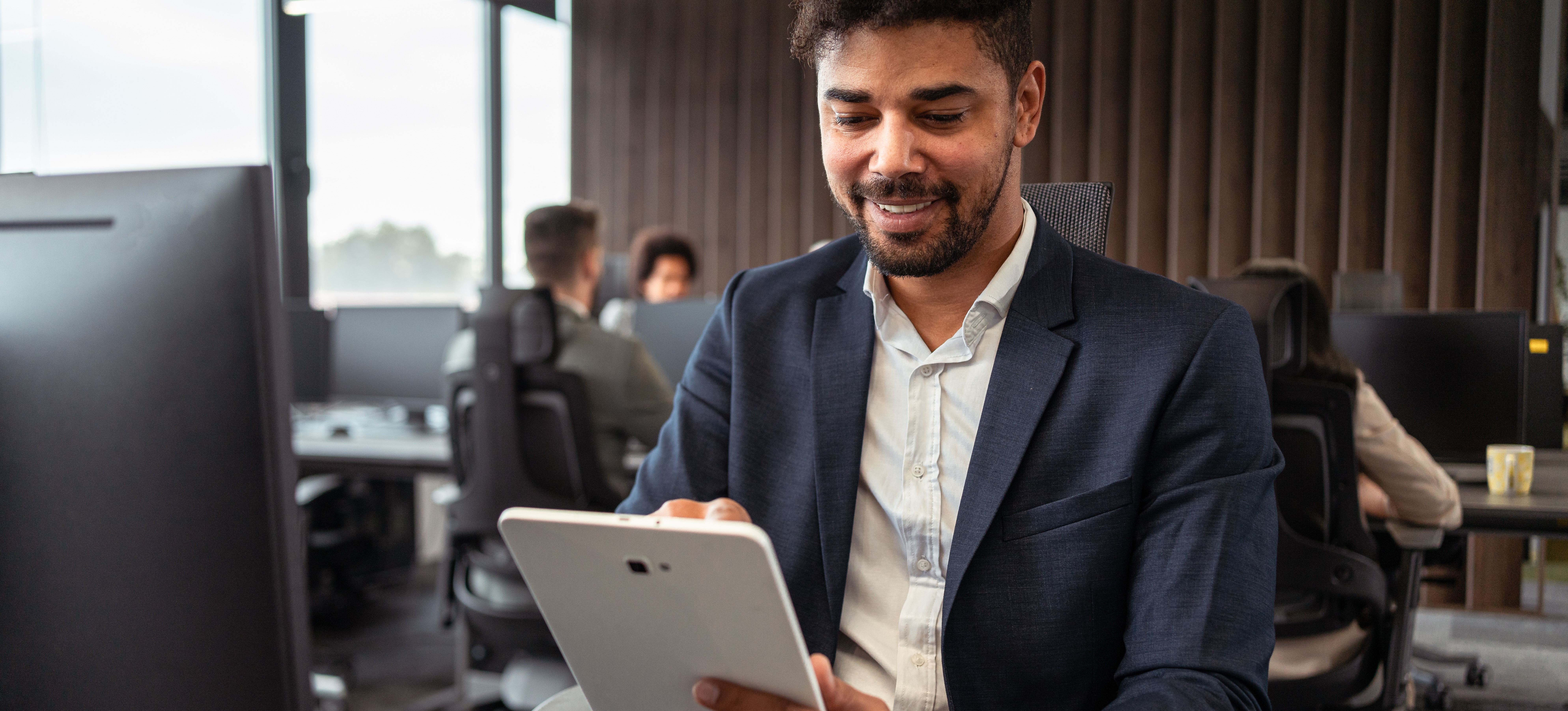 [Featured image] A professional who works in a computer science job uses a tablet while sitting at an office desk in front of a computer, with colleagues at their workstations in the background.
