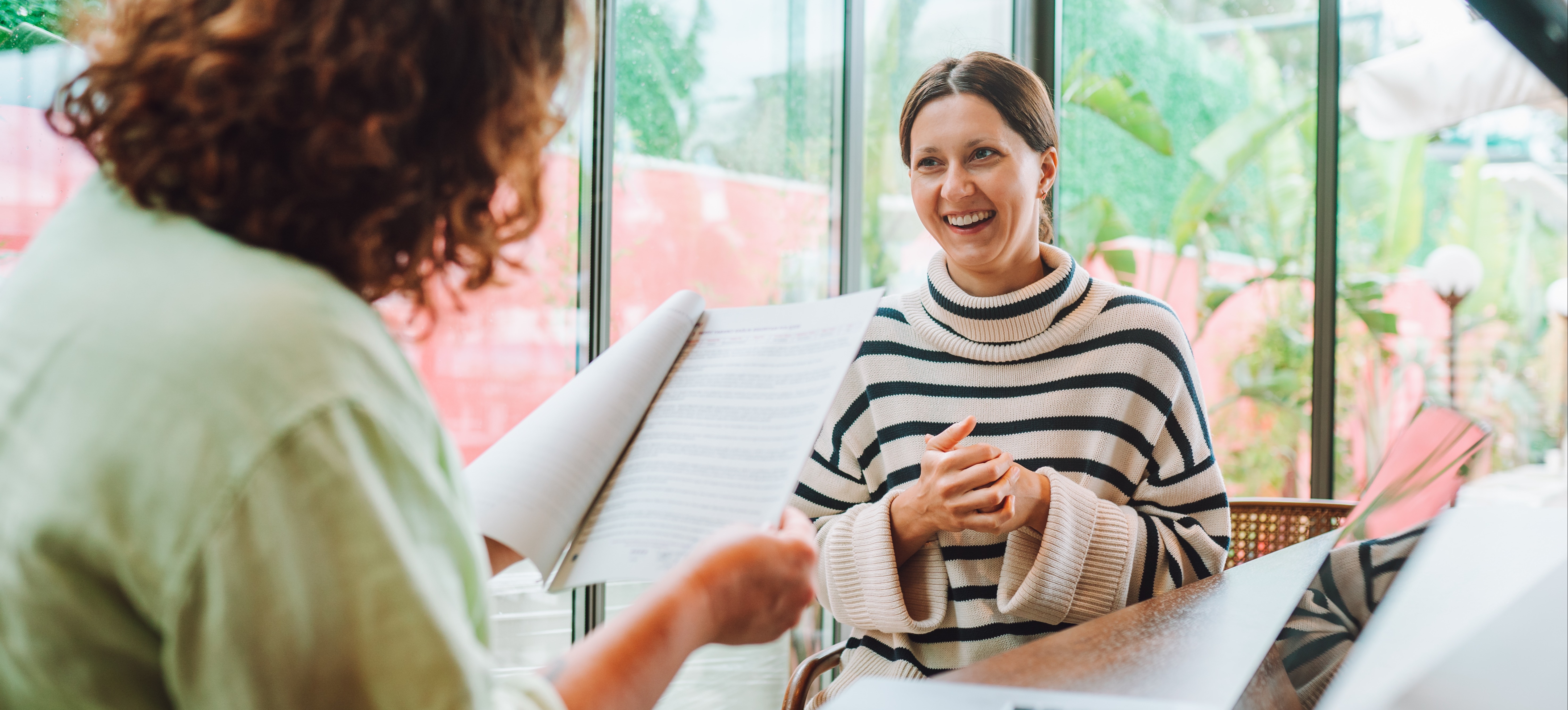 [Featured Image] Two females sit at a desk and discuss teacher interview questions for the new job.