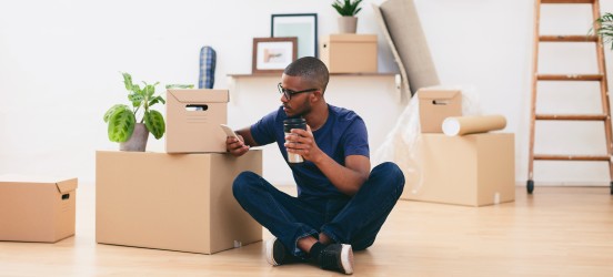[Image en vedette] Un homme consulte son téléphone dans son nouveau logement à la recherche d'offres d'emploi après avoir déménagé sans travailler.
