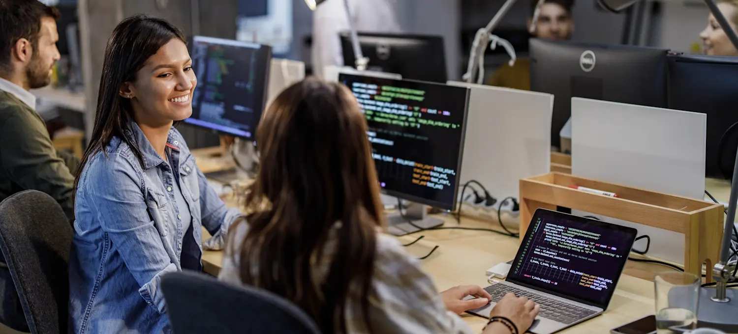 [Featured Image] DevOps professionals interact with each other in a workspace while discussing the best way to learn Docker and viewing code on their computers.