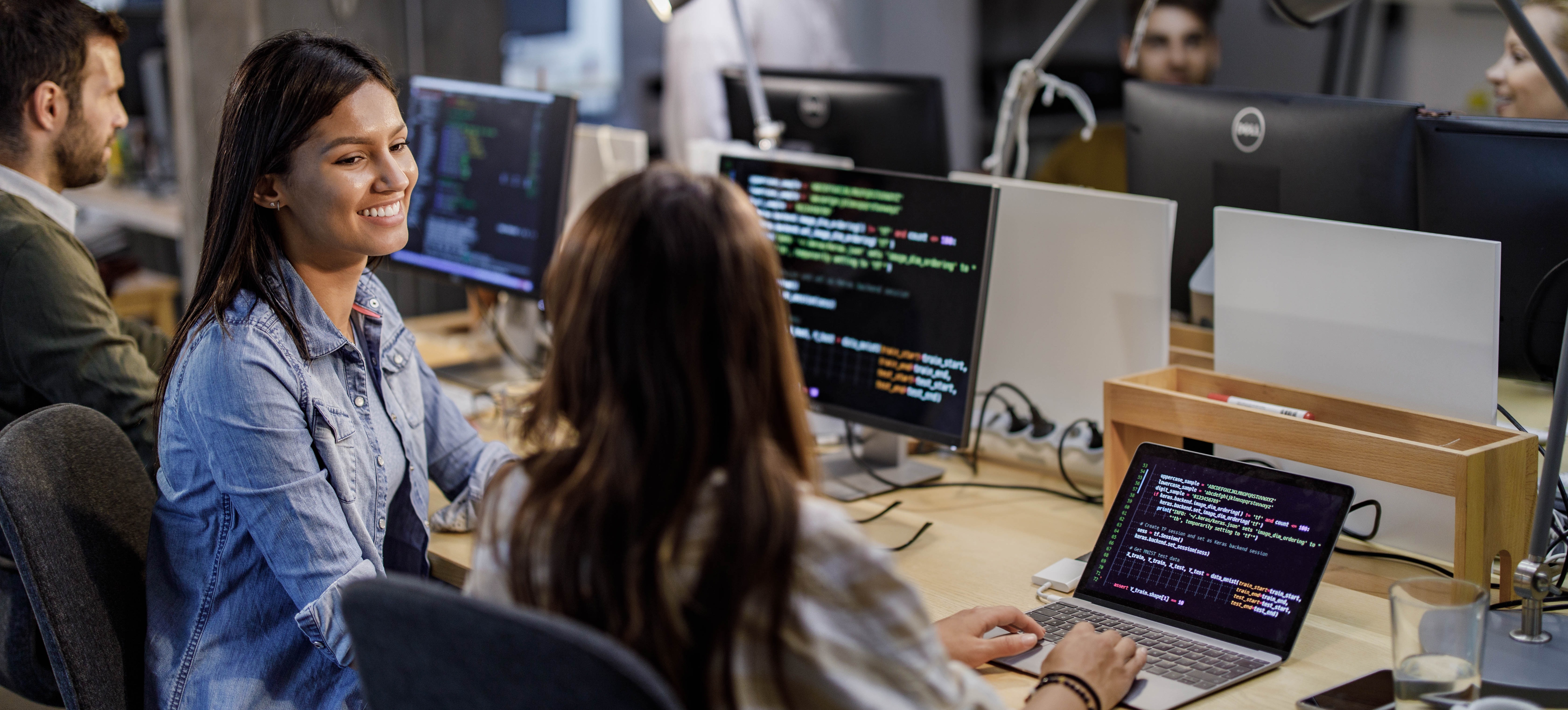 [Featured Image] DevOps professionals interact with each other in a workspace while discussing the best way to learn Docker and viewing code on their computers.
