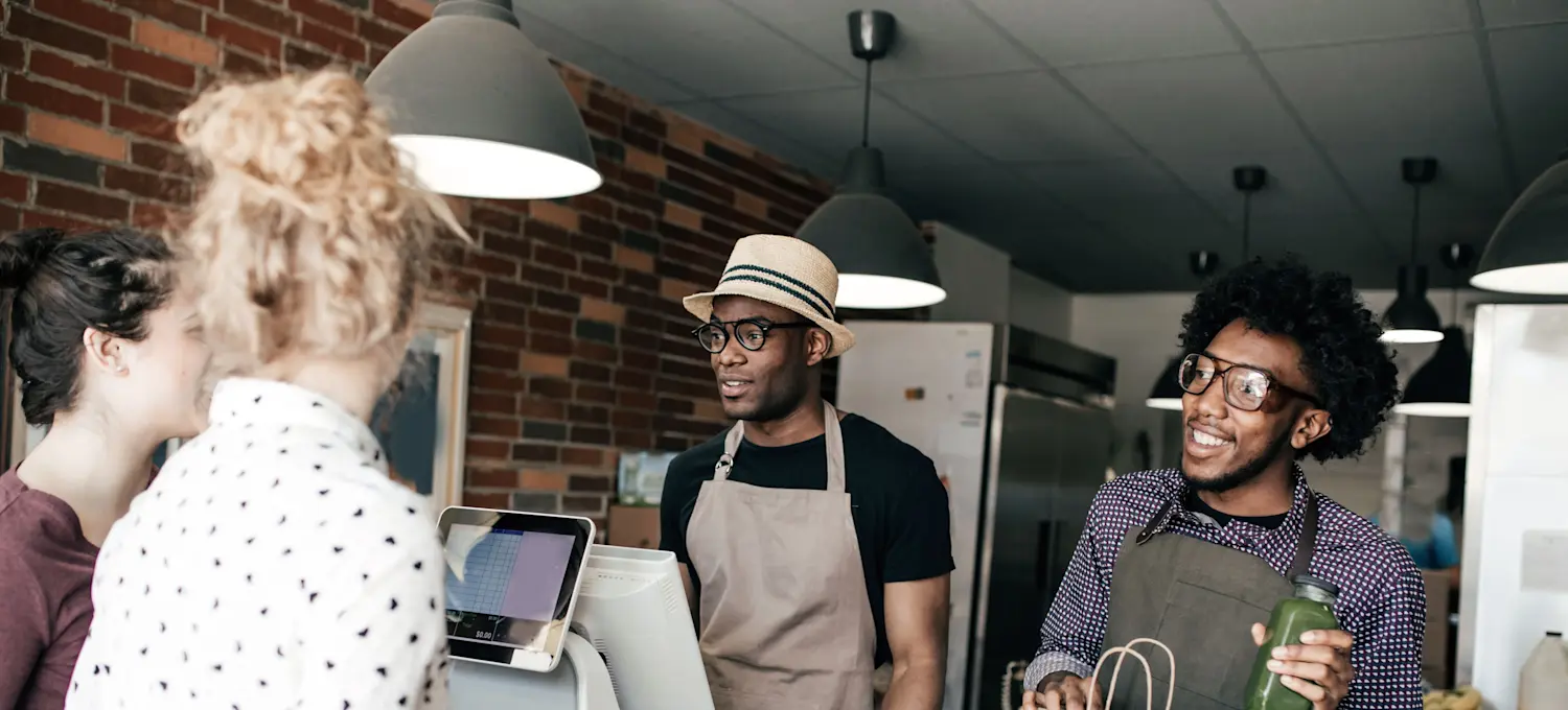 [Featured Image]  Two people, both wearing glasses and aprons, serve their customers at their business.