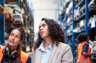 [Featured image] Two product managers are wearing an orange vest, and gloves looking at warehouse products. 