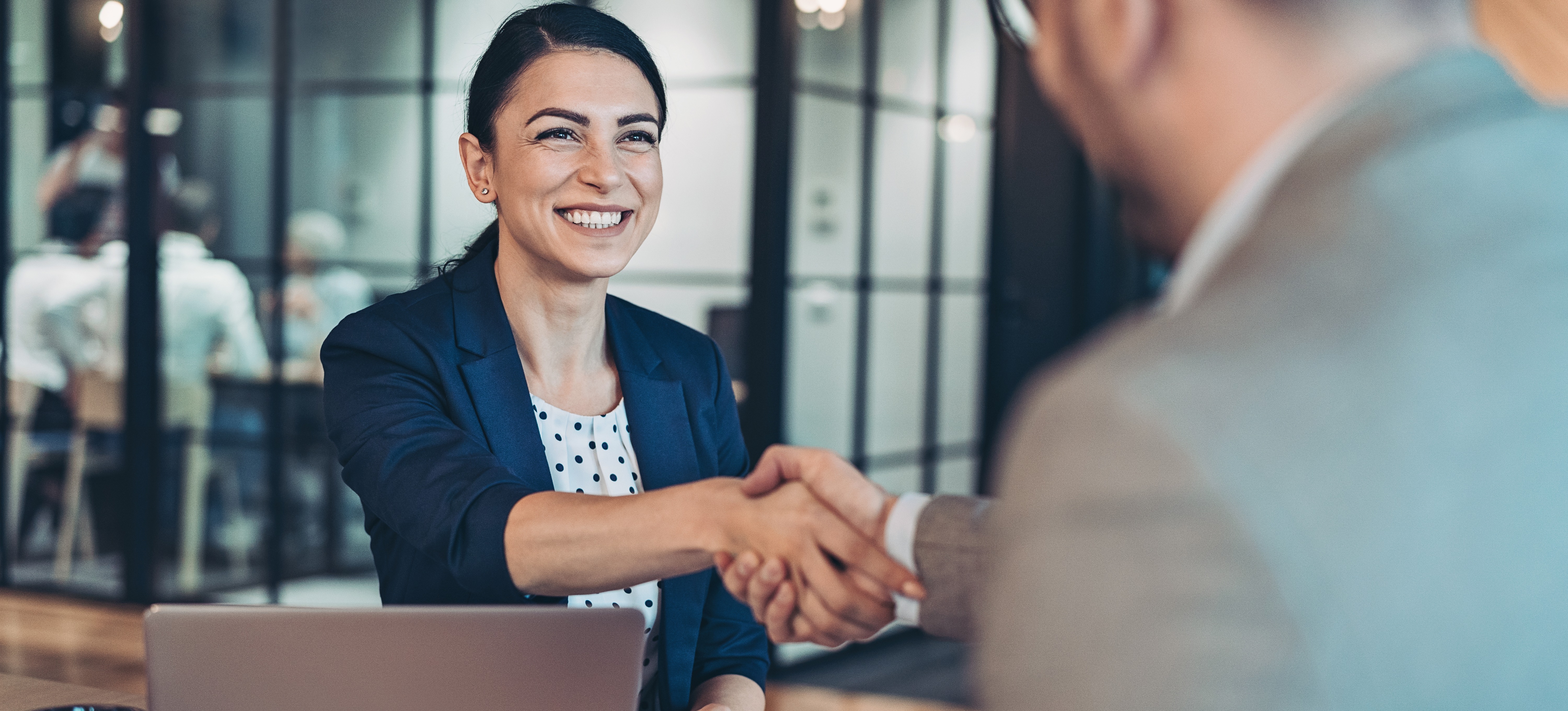 [Featured Image] In an office setting, a woman interviewer, prepared to ask supply chain analyst interview questions, shakes her interviewee's hand ahead of an interview for a supply chain analyst job.
