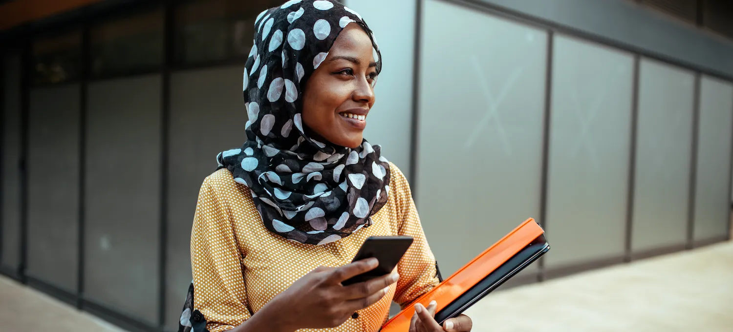 [Featured image] A communications degree student walks on campus with their phone and a folder of coursework.