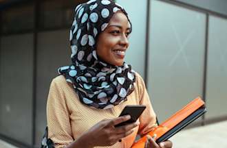 [Featured image] A communications degree student walks on campus with her phone and a folder of coursework.