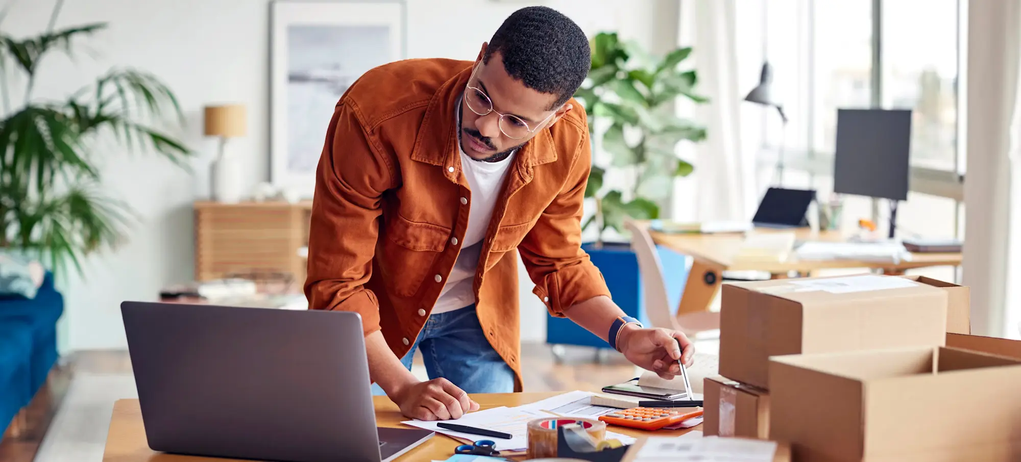 [Featured image] A person packs boxes for their ecommerce business from home and leans over table doing calculations on the calculator.