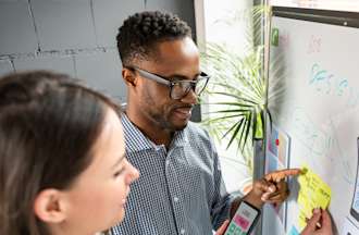 [Featured image] A male, wearing a blue shirt and glasses, and a female wearing a white and red top are working on a whiteboard with charts and notes as they discuss game development. 