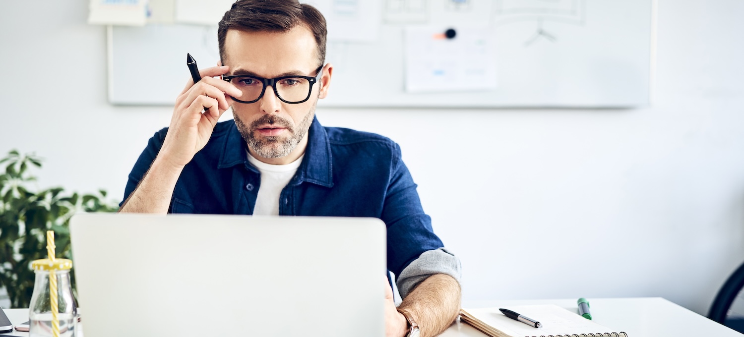 [Featured image] A person wearing glasses and holding a pen does research on his laptop about earning an MBA with a low GPA.