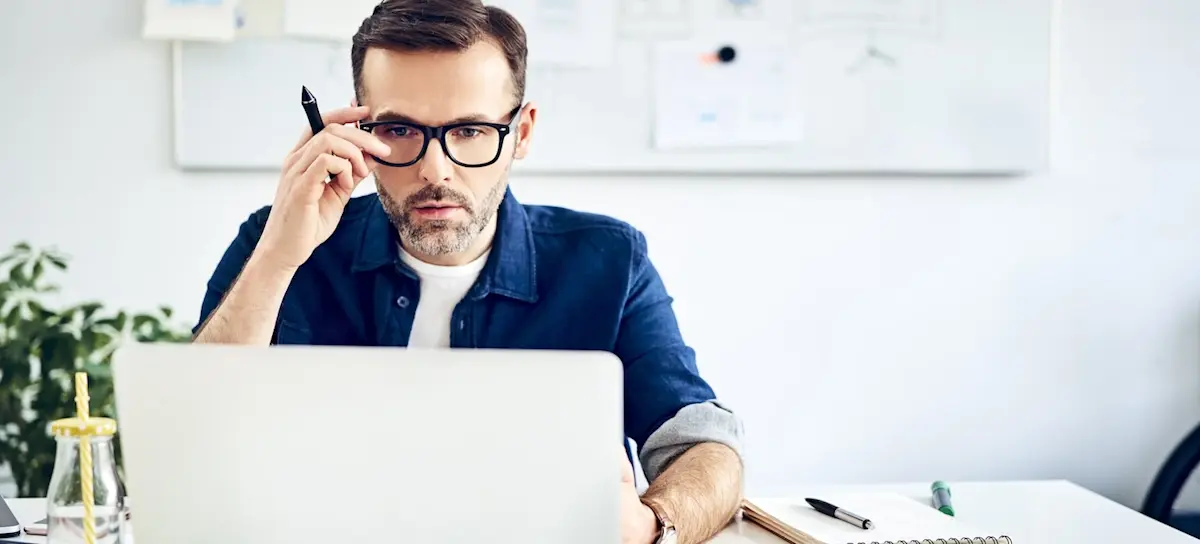 [Featured image] A person wearing glasses and holding a pen does research on his laptop about earning an MBA with a low GPA.