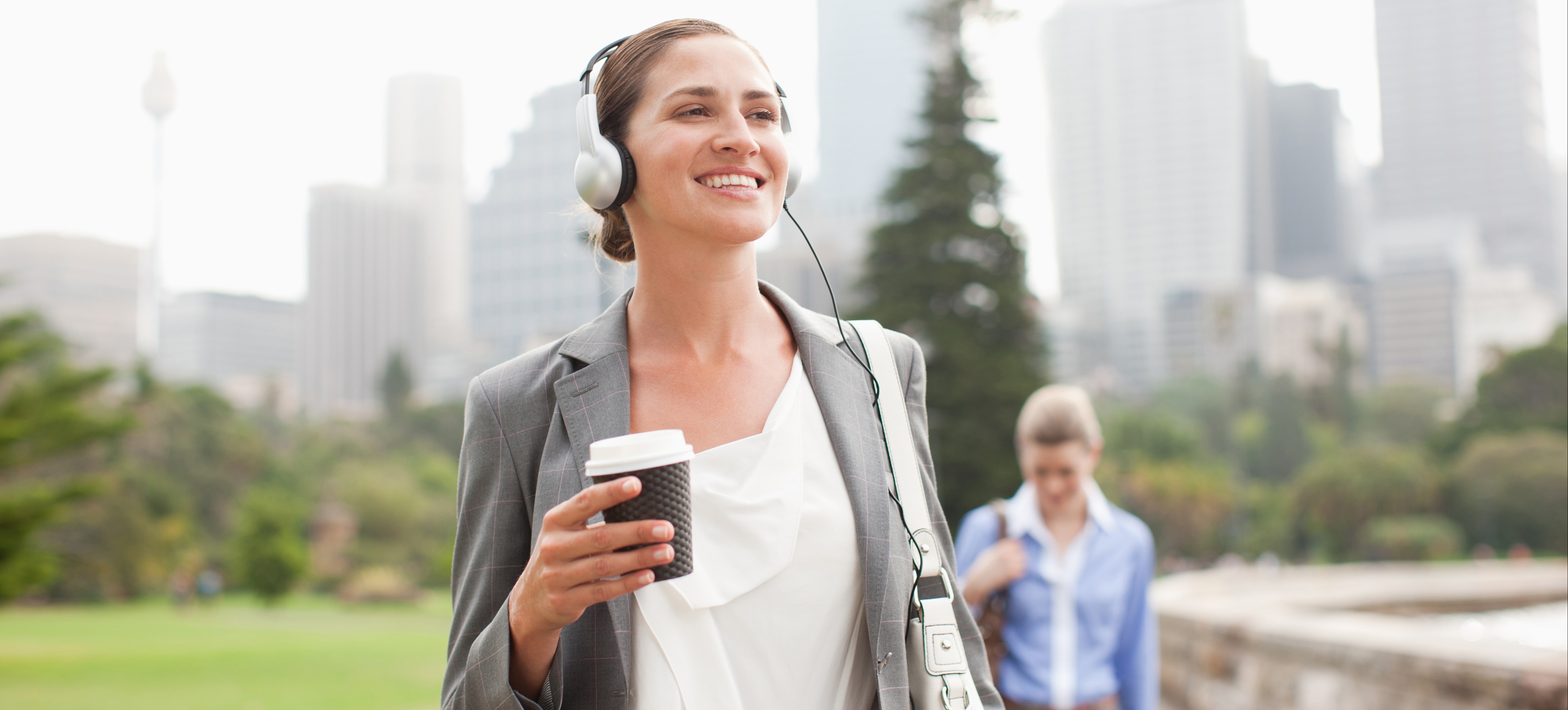 [Featured Image] A woman holding coffee and listening to headphones walks to work in one of the best cities for cybersecurity jobs.